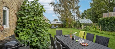 Une terrasse avec une table et des chaises, un barbecue et une grande plante à feuillage abondant.