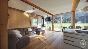 A spacious living room with a wooden ceiling, a grey sofa, a white coffee table, and a kitchen area with a large window. - Premium Chalet # 8 with sauna and swim spa (Murau)