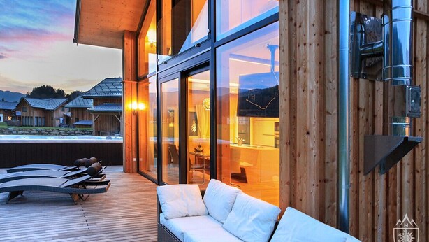 A modern outdoor seating area with lounge chairs and a sofa, large glass windows, and a wooden deck. - Premium Chalet # 8 with sauna and swim spa (Murau)