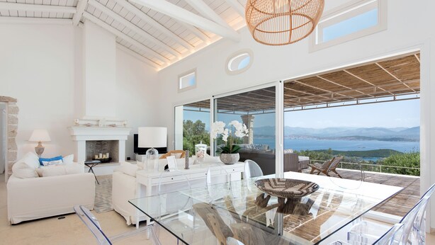 A spacious living room with a glass table, white furniture, and a view of the sea. - Thea Lofou House (Corfu)