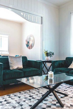 Un salon moderne avec une table basse en verre, deux canapés verts, un miroir rond et une plante en pot.