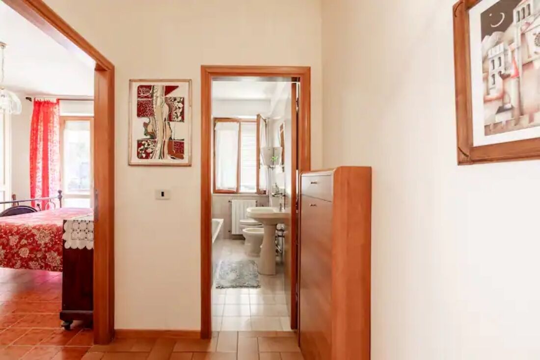 Un pasillo que conduce a un baño con lavamanos e inodoro, una cama con colcha estampada y un cuadro en la pared.