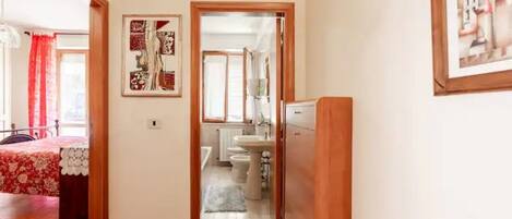 Un pasillo que conduce a un baño con lavamanos e inodoro, una cama con colcha estampada y un cuadro en la pared.