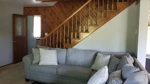 Un salon avec un canapé gris, un escalier en bois et une porte dotée d’un panneau en verre dépoli.