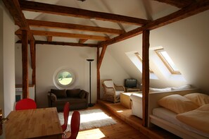 A cozy attic room with a wooden ceiling, a round window, a sofa, a TV, and a bed.