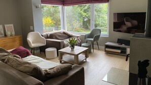 Ein Wohnzimmer mit Sofa, Sesseln, einem Couchtisch und einem Fernseher. Große Fenster mit Vorhängen, eine Holzkommode und ein kleiner Tisch mit einer Vase sind ebenfalls vorhanden.