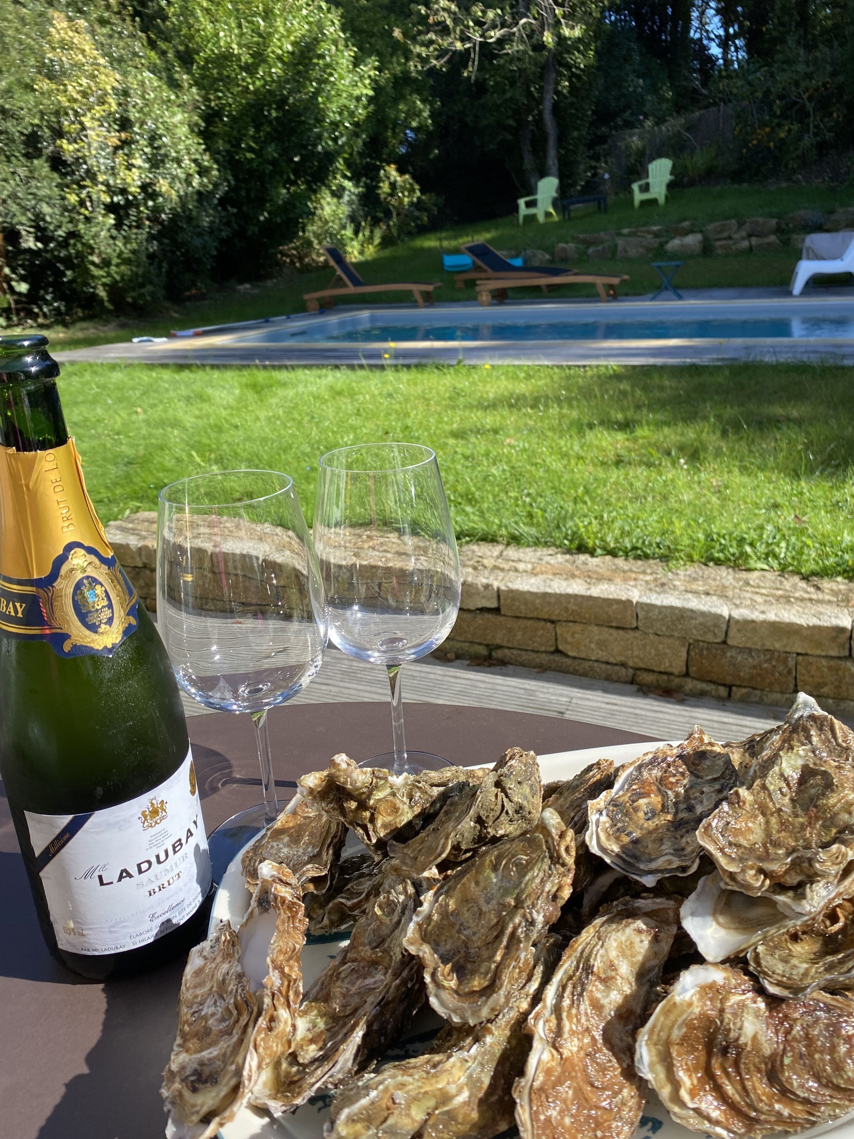 Eine Flasche Brut de Ladur Champagner, zwei leere Gläser und eine Platte Austern auf einem Tisch mit Blick auf eine Wiese und eine Straße im Hintergrund.