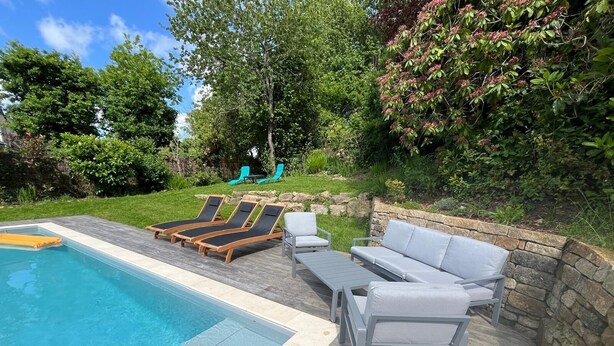 Ein Swimmingpool mit Liegestühlen und einer Sitzecke, umgeben von Grünflächen und einer Steinmauer.