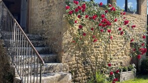 Un bâtiment en pierre avec un escalier menant à une porte, le tout entouré d’un jardin où poussent des fleurs rouges.