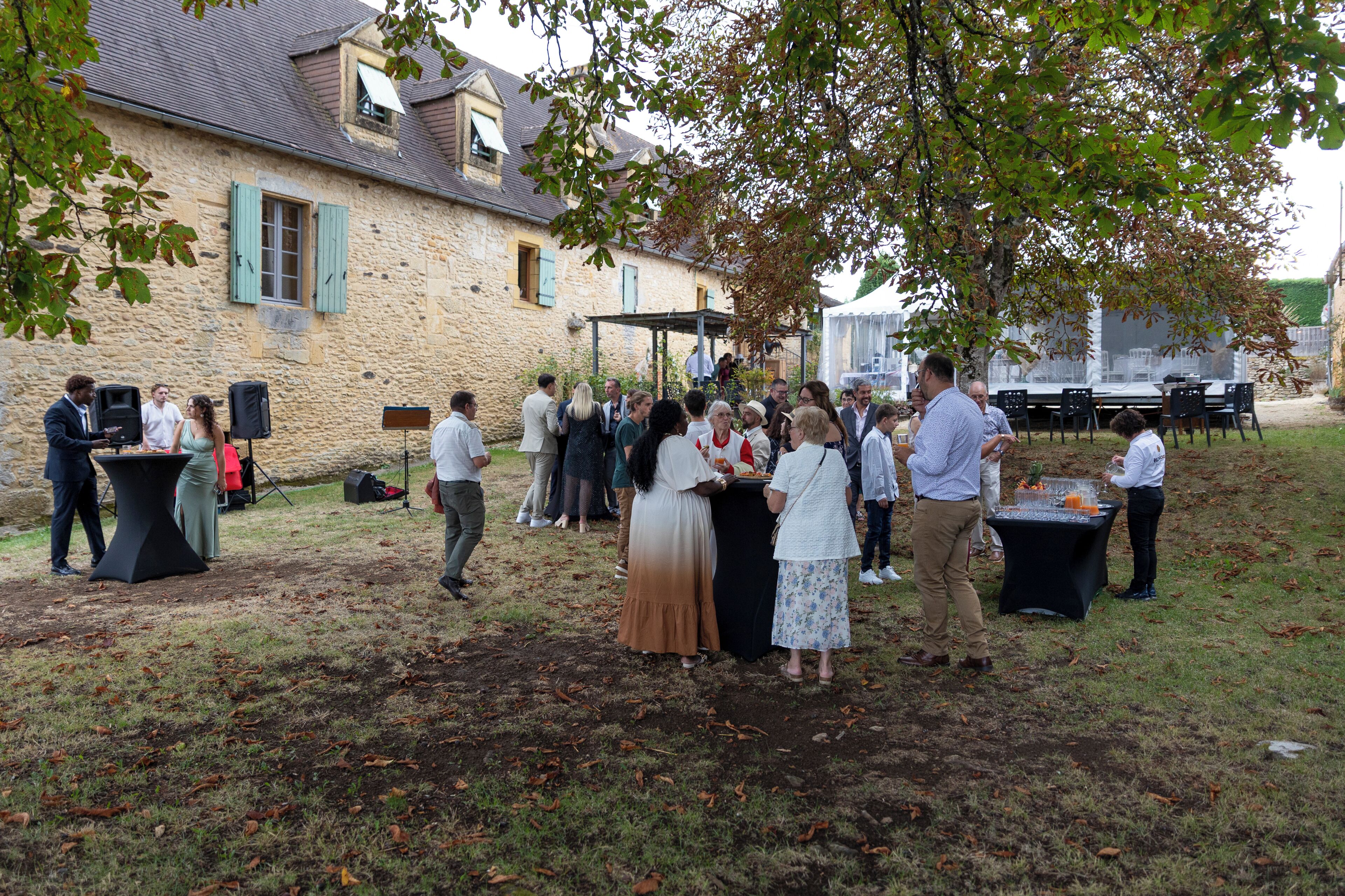 A stone building with a covered outdoor area, a table with a speaker, and a group of people gathered.