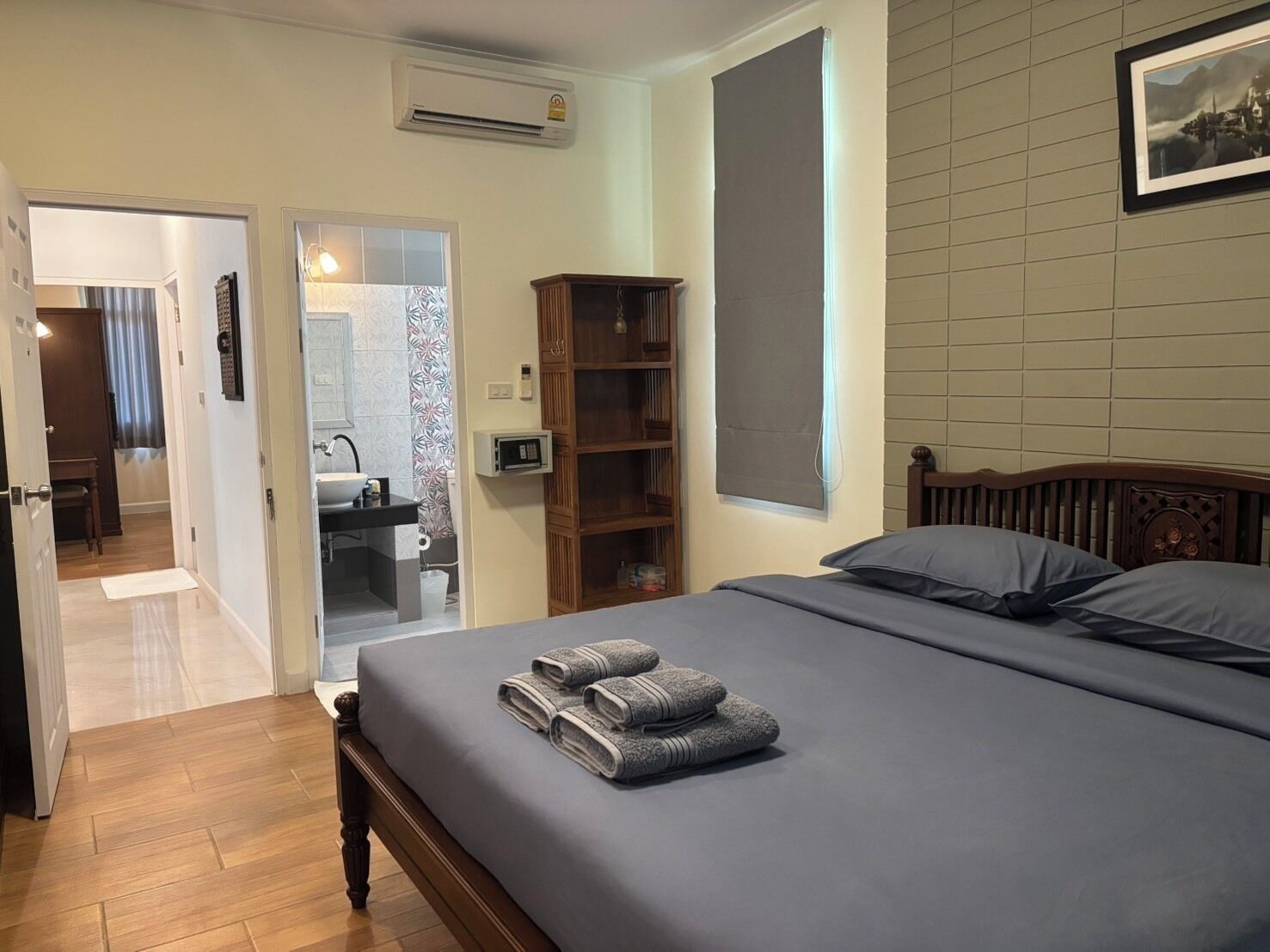 Un dormitorio con cama, cabecera de madera, mesita de noche, aire acondicionado instalado en la pared, estante con libros y un cuadro enmarcado.