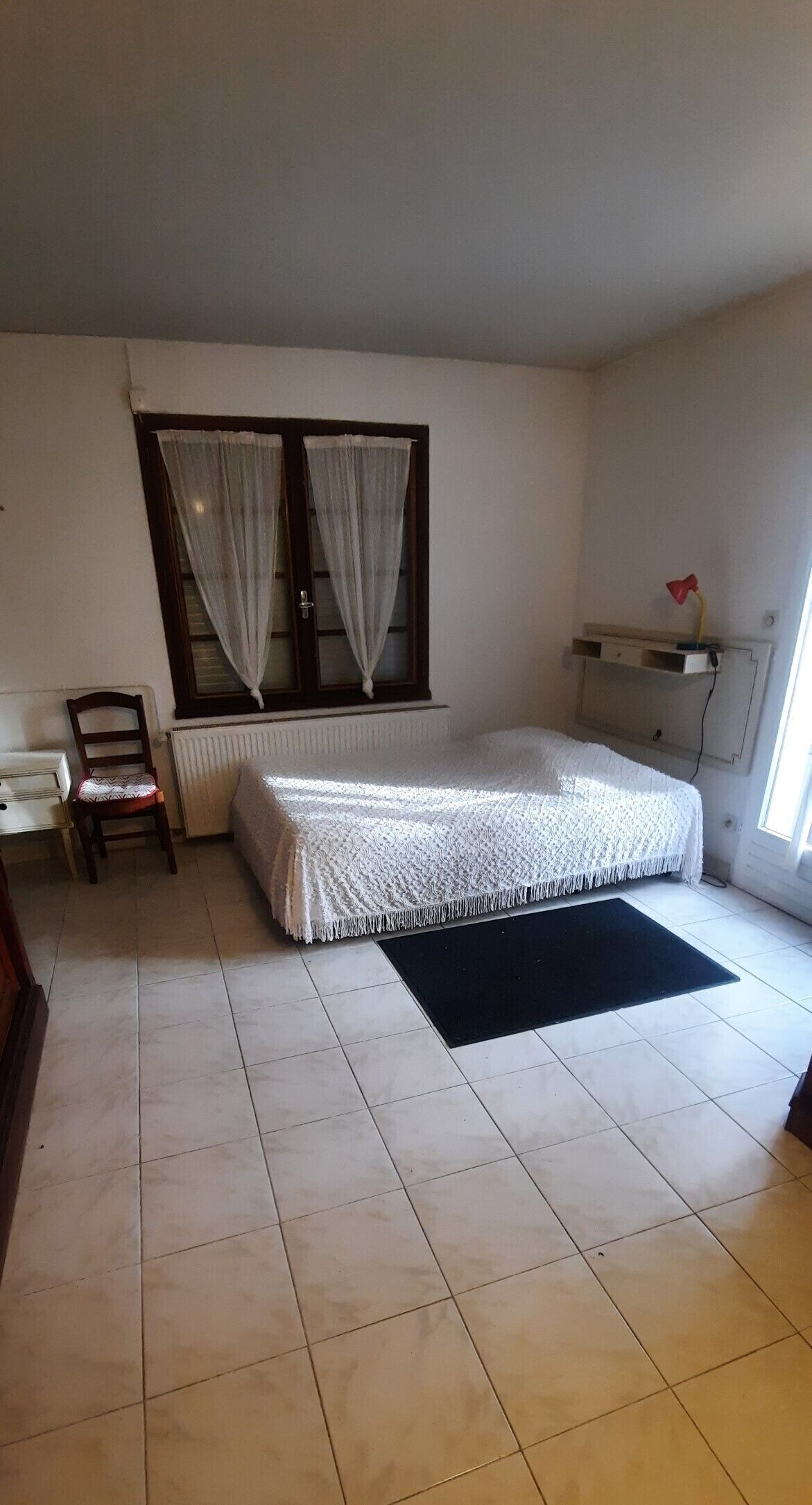 Une chambre à coucher avec un lit, une chaise, un radiateur et une fenêtre avec des rideaux.