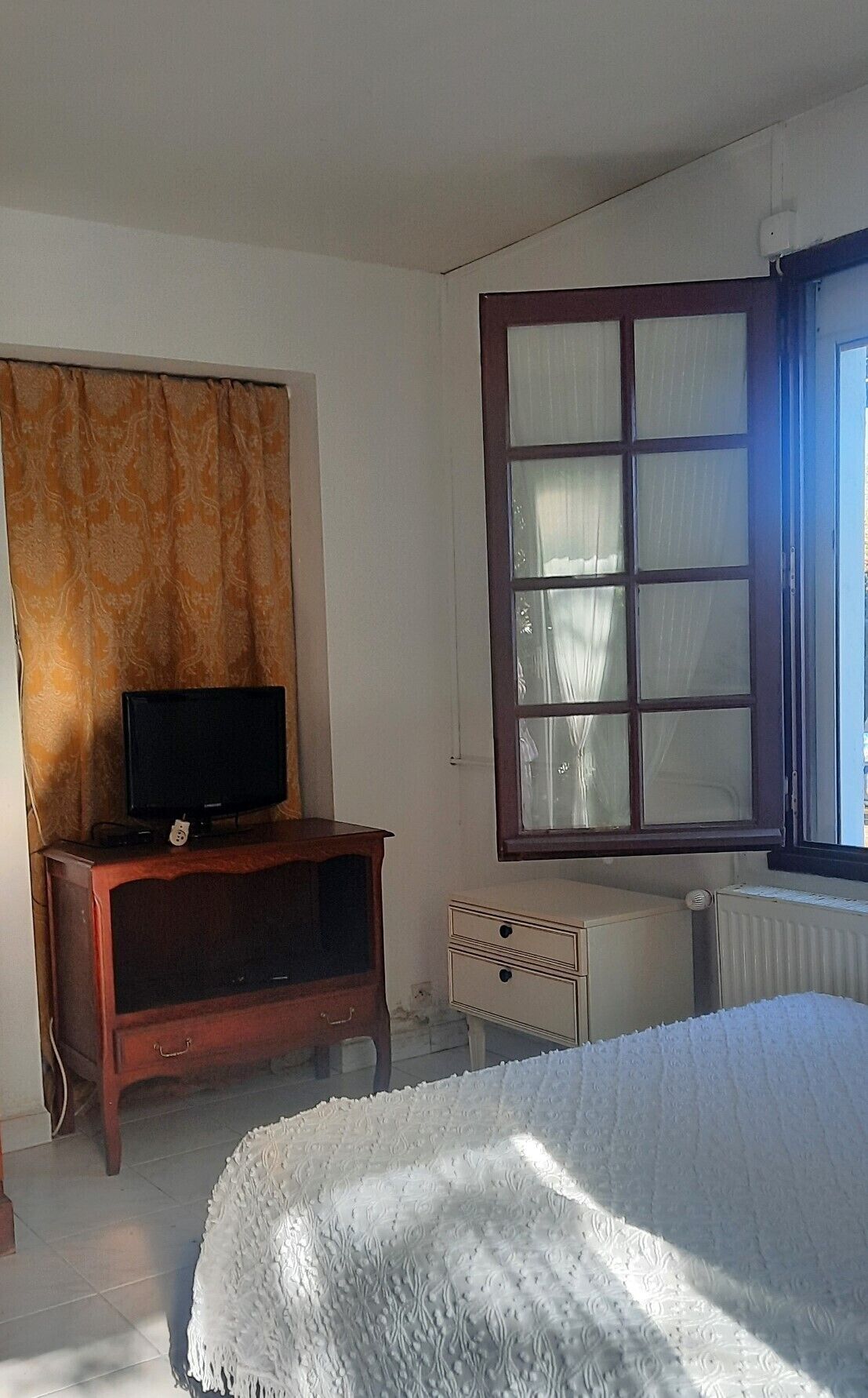 Une chambre à coucher avec un lit, un téléviseur posé sur un meuble en bois, un radiateur et une fenêtre avec des rideaux.