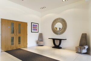 Un vestíbulo moderno con puerta de madera, espejo redondo, un cuadro enmarcado y dos sillones.