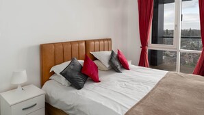 A neatly made bed with a brown headboard, white bedding, and several pillows in various colors. A white bedside table with a lamp and a drawer is visible to the left. The room has a large window with red curtains, offering a view of buildings outside. - KD Tower Abodebed (Hemel Hempstead)