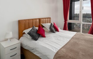 Una cama cuidadosamente tendida con un cabecero marrón, ropa de cama blanca y varios cojines de diferentes colores. A la izquierda, una mesita de noche blanca con una lámpara y un cajón. La habitación tiene un ventanal grande con cortinas rojas, que ofrece vistas de edificios exteriores.