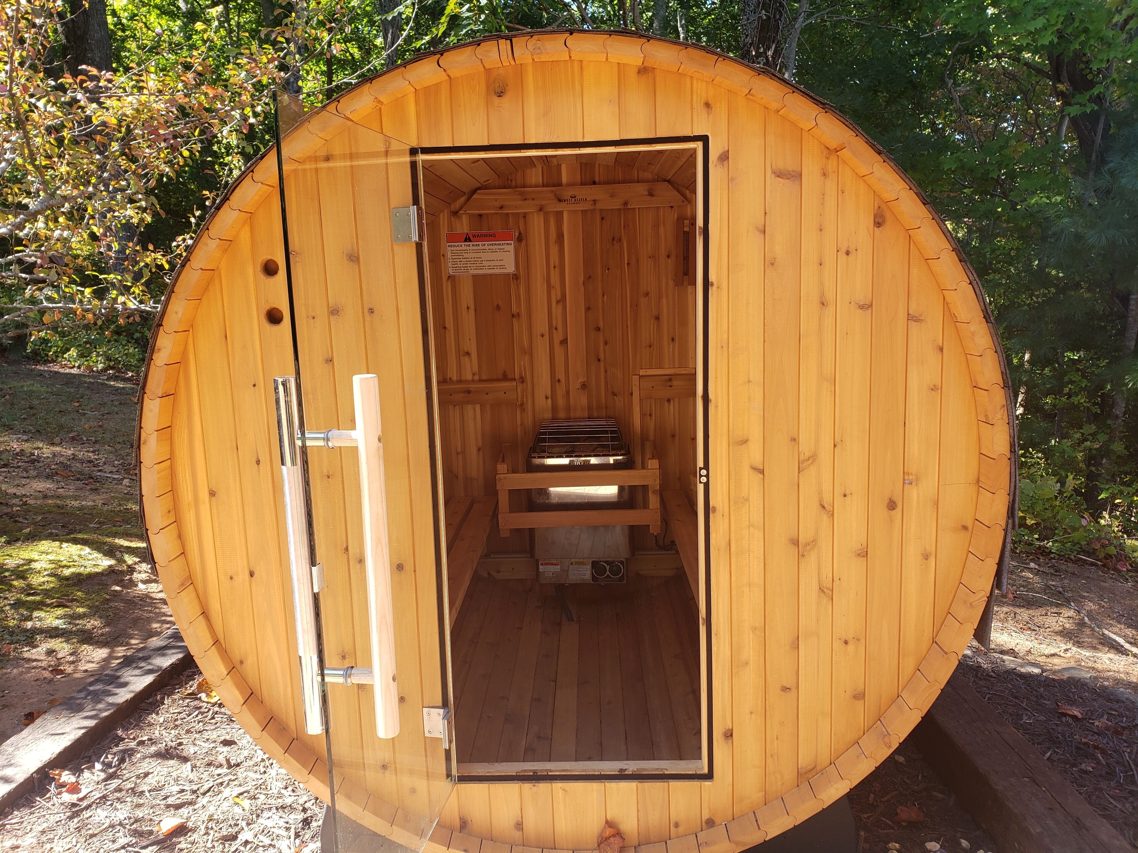 Eine runde Sauna aus Holz mit Tür, einer Sitzbank darin und einem Warnschild.