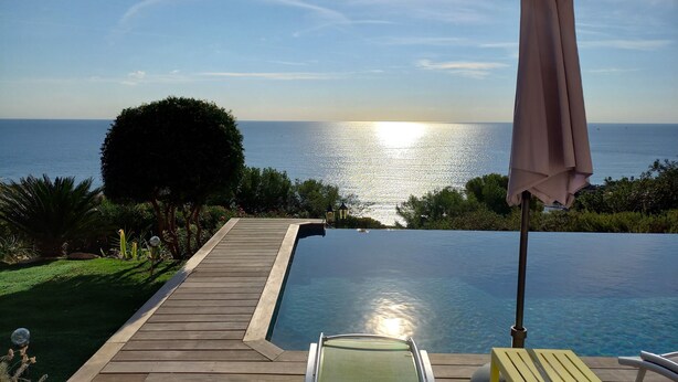 Eine Holzterrasse führt zu einem klaren Pool mit Blick auf das Meer und einen Sonnenuntergang.