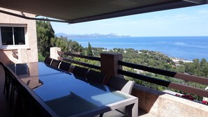 Ein Balkon mit Glastisch und Stühlen, der Blick auf eine Küstenlandschaft mit Bergen und dem Meer bietet.