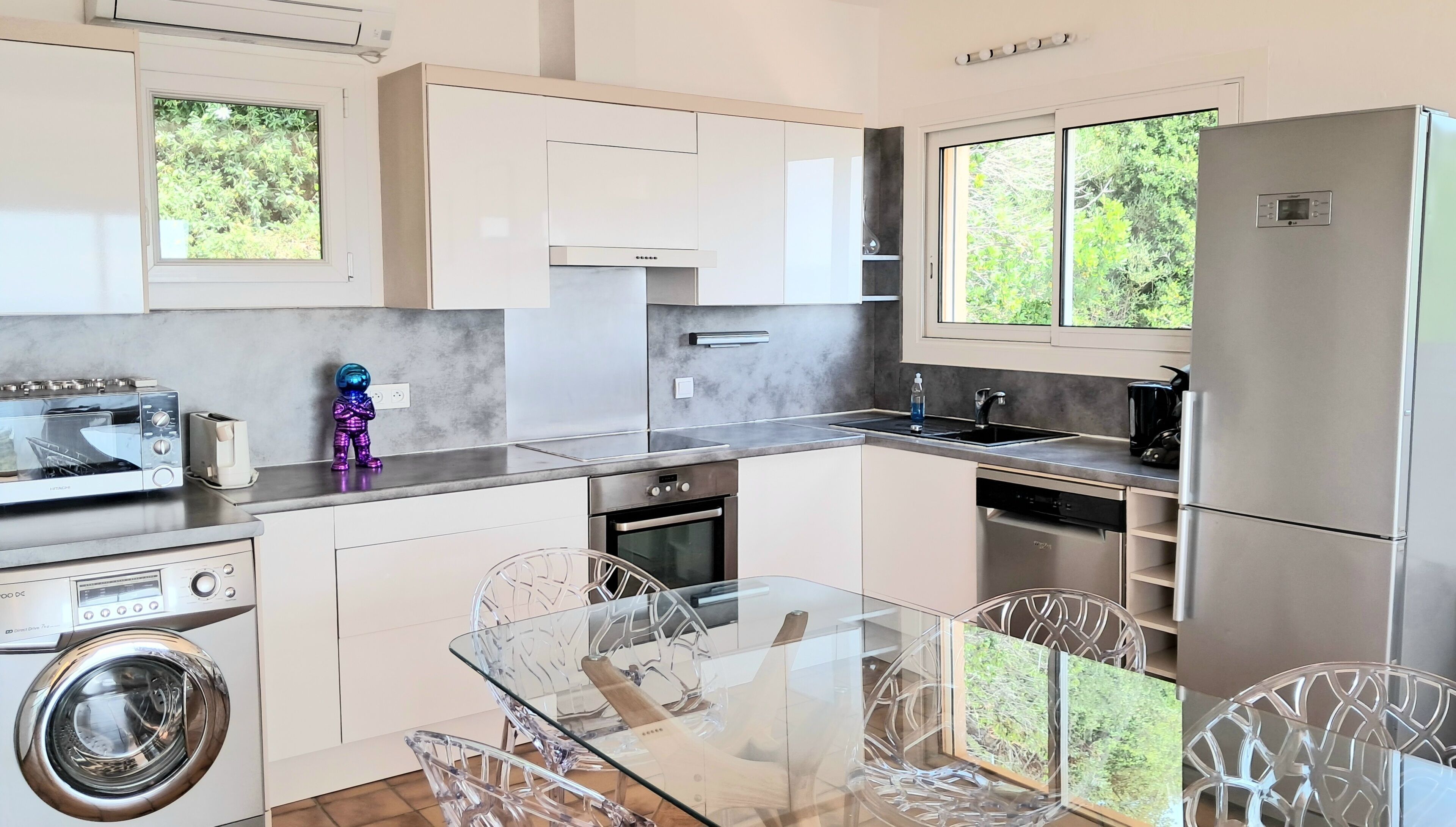 A modern kitchen with stainless steel appliances, a glass dining table, and a view of greenery outside.