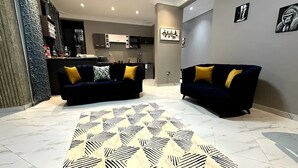 A modern living room with two dark sofas and patterned rugs, a kitchen in the background, and artwork on the walls.