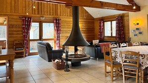 A cozy log cabin interior with a central wood-burning stove, wooden beams, a dining area, and a seating space. - WONDERFUL SPACIOUS CHALET IN BOURG D’OISANS - SLEEPS 10(*supplement available up to 16) (Le Bourg-d'Oisans)