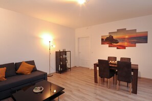 A living room with a sofa, coffee table, dining table with chairs, a bookshelf, and a wall-mounted artwork.