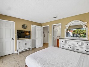 A bedroom with a large bed, a dresser, a television, and a door leading to another room.