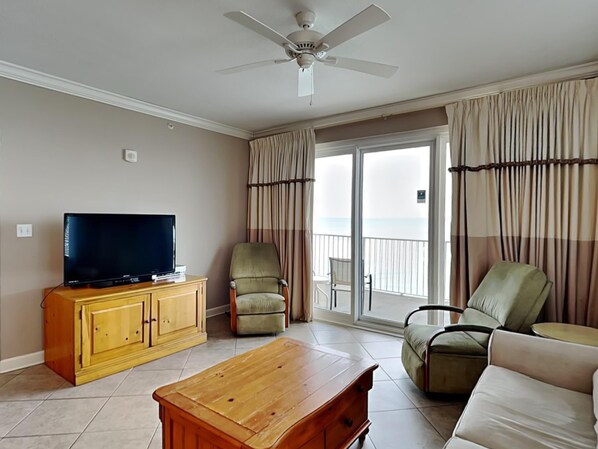 A living room with a television on a wooden cabinet, two chairs, a sofa, and a ceiling fan. - Grandview East #1005 | Near Area Fun! (Panama City Beach)
