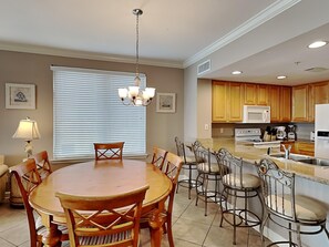 A dining area with a round wooden table and chairs, a chandelier, and a window with blinds. - Grandview East #1005 | Near Area Fun! (Panama City Beach)