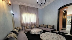 Ein Wohnzimmer mit einer Sofagarnitur, zwei runden Tischen mit Spitzenbettwäsche, einer Chandelierleuchtre und Blick ins Freie.