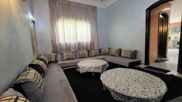 A living room with a sofa set, two round tables with lace tablecloths, a chandelier, and a view of the outside.