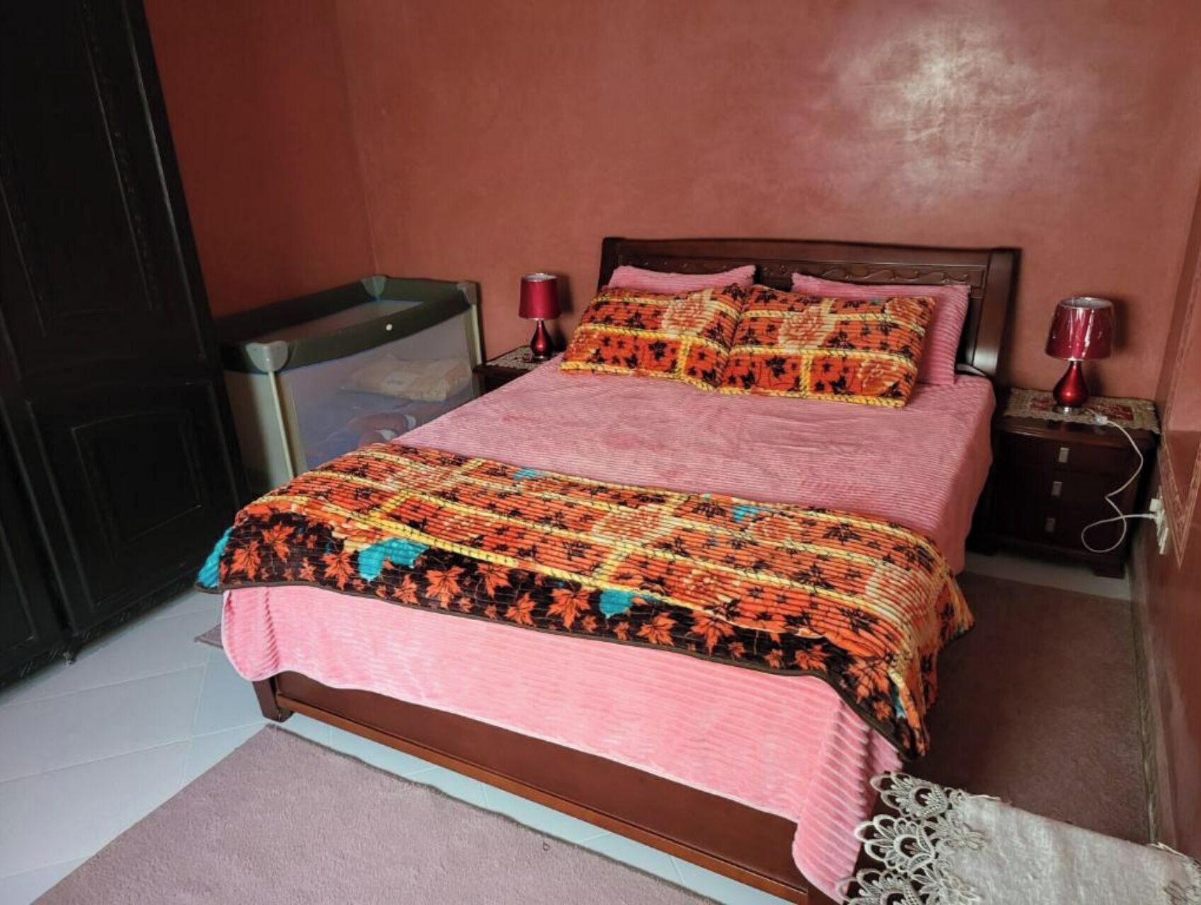 A bed with a patterned bedspread and pillows, a bedside table with a lamp, and a window with a view of the outside.