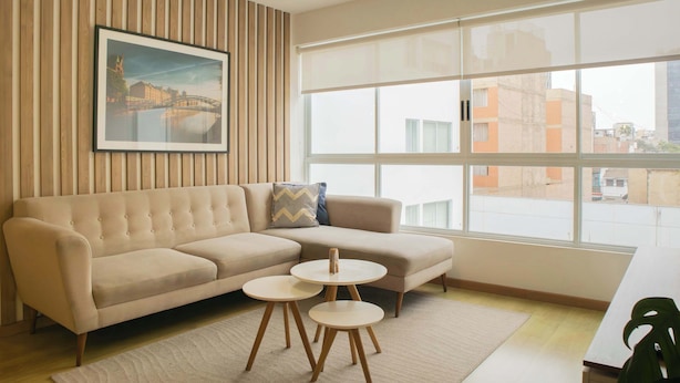 Una sala de estar moderna con un sofá seccional color beige, dos mesas de centro redondas, un cuadro enmarco con una vista urbana, y un ventanal con vistas a edificios.