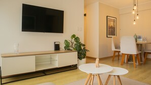 Una sala moderna con un televisor de pantalla plana, una consola de madera, una planta en maceta, un comedor con sillas y un lámpara colgante.