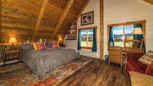 A cozy bedroom with a wooden ceiling, a large bed with a patterned rug, a dresser, and a view of the outdoors through a window.