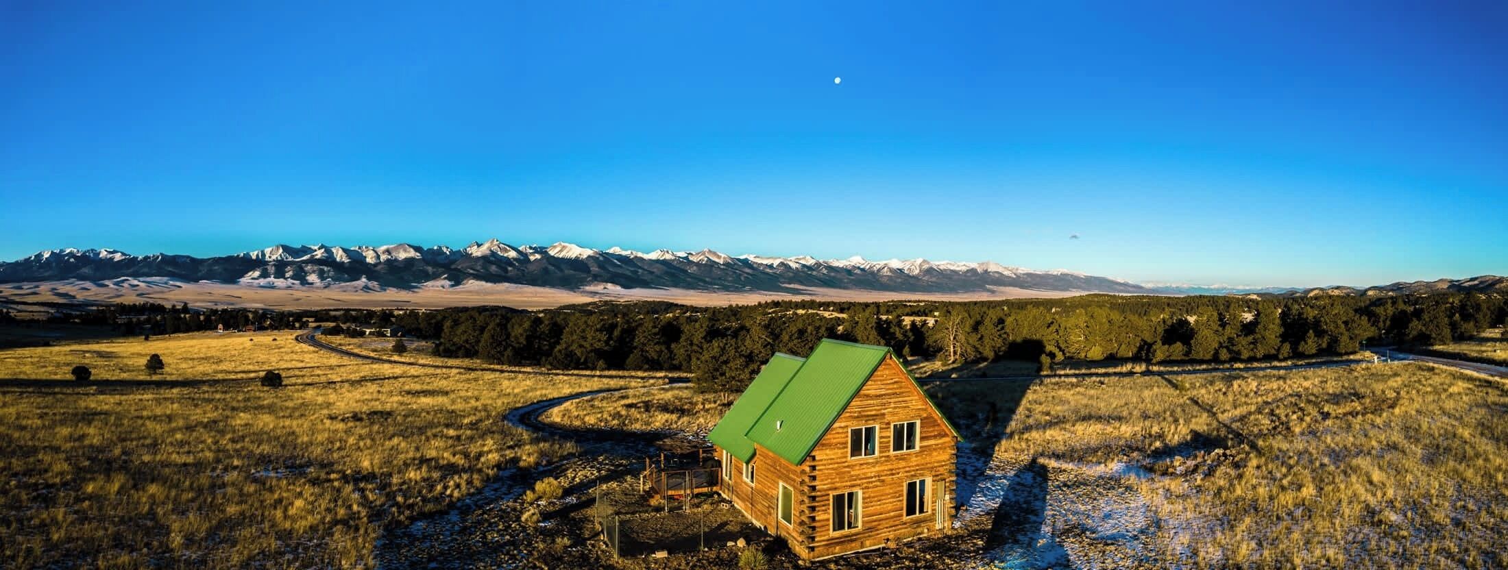 Eine Holzhütte mit einem grünen Dach, umgeben von einem grasbewachsen Feld und einem Bergpanorama im Hintergrund.