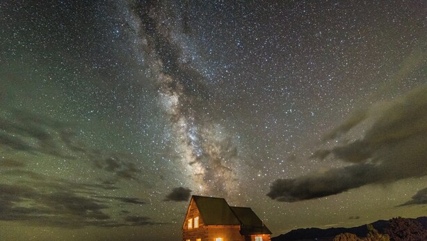 A log cabin with a single window and a door, surrounded by a field under a starry night sky.