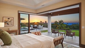 A bedroom with a large bed, a view of a patio with outdoor furniture, and a sunset view through the glass doors. - Hula House: Luxury Villa w/ Superb Views/Amenities (Koloa)