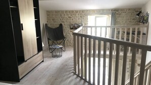 A loft-style room with a high ceiling, exposed stone walls, a wooden floor, a white railing, and a black chair with a magazine. - A peaceful, restful little corner (Annezay)