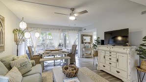 A living room with a sofa, coffee table, and TV. There is a ceiling fan, a dining table with chairs, and a white cabinet with drawers. - Sunset Rentals | Treetops 5701 | Dog Friendly (Hilton Head Island)