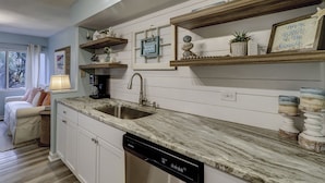 A modern kitchen with a built-in dishwasher, wooden shelves with decorative items, and a marble countertop. - Sunset Rentals | Treetops 5701 | Dog Friendly (Hilton Head Island)