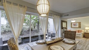 A dining area with a table set for four, large windows with curtains, a hanging light fixture, and a wicker chair. - Sunset Rentals | Treetops 5701 | Dog Friendly (Hilton Head Island)