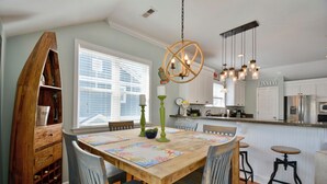 Een eetruimte met een houten tafel, stoelen en een kroonluchter. In de achtergrond is een keuken te zien met witte kasten en een koelkast.