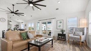 A spacious living room with a leather sofa, a coffee table, and a patterned armchair. There are ceiling fans, a large window with a view, and a kitchen in the background.