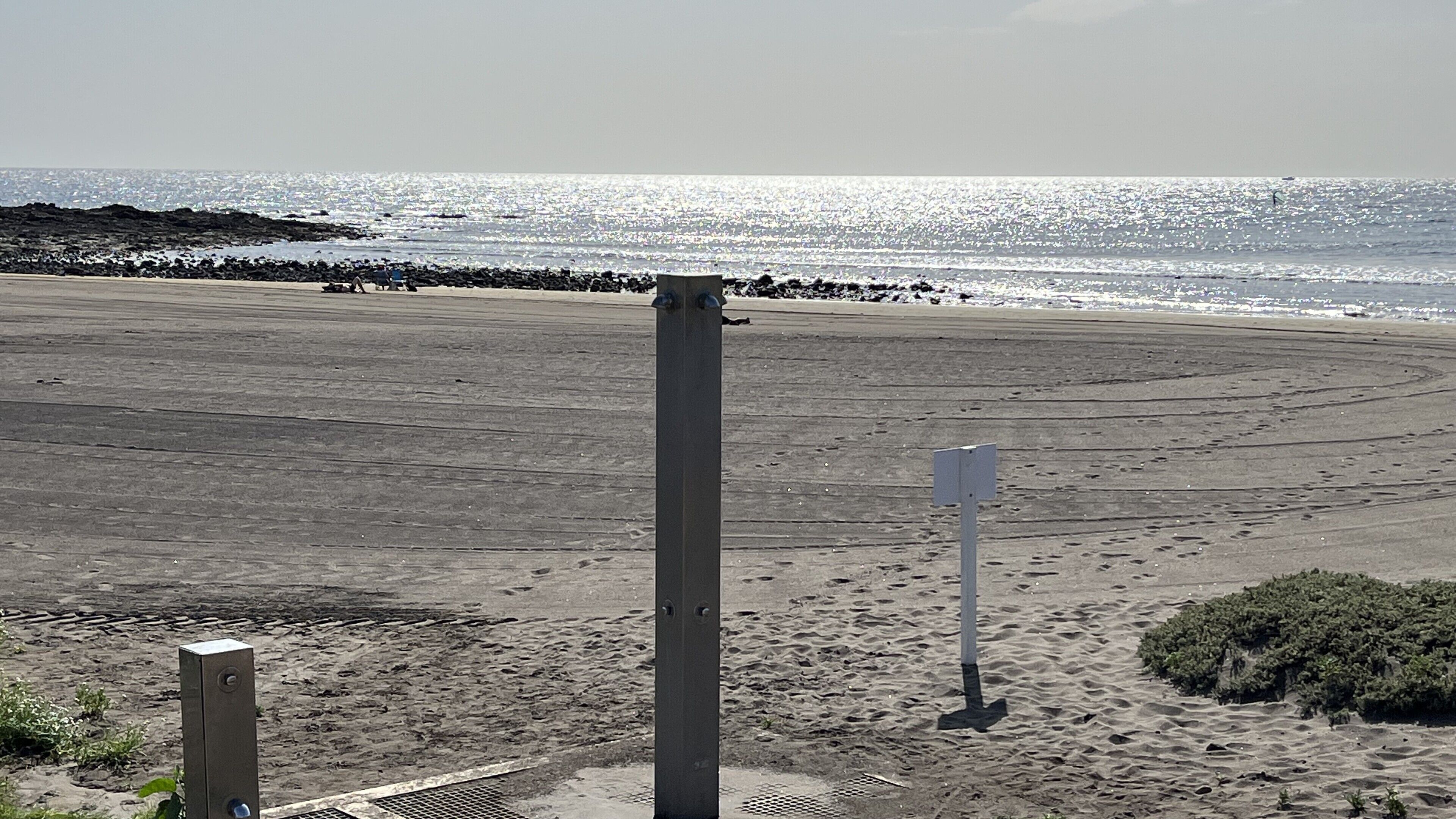 Ein Strand mit einem Metallpfahl im Vordergrund, einer felsigen Küstenlinie und freiem Blick auf den Ozean.