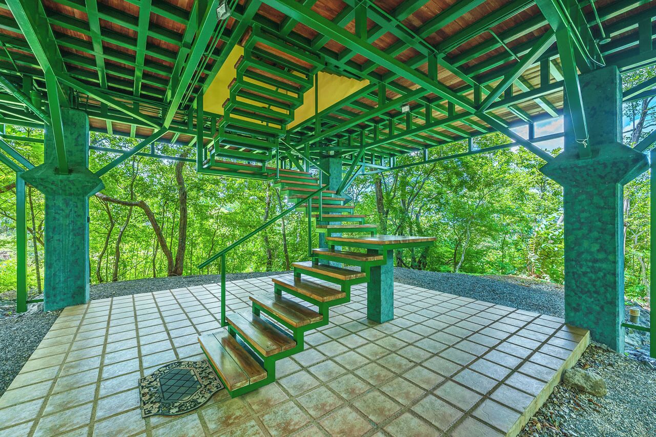 A spiral staircase with green railings and wooden steps, leading to a covered area with a tiled floor and a view of trees.
