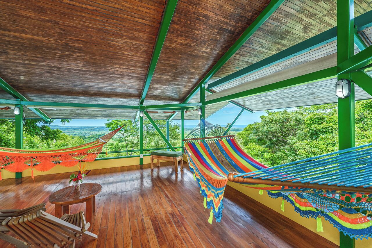A hammock with a colorful woven pattern, a wooden table, and a view of lush greenery through large windows.