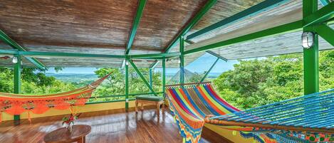 A hammock with a colorful woven pattern, a wooden table, and a view of lush greenery through large windows.