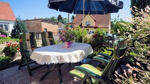 Ein runder Tisch mit weißer Tischdecke, zwei Stühle mit grün-beiger Polsterung und ein großer blauer Sonnenschirm, der Schatten spendet.